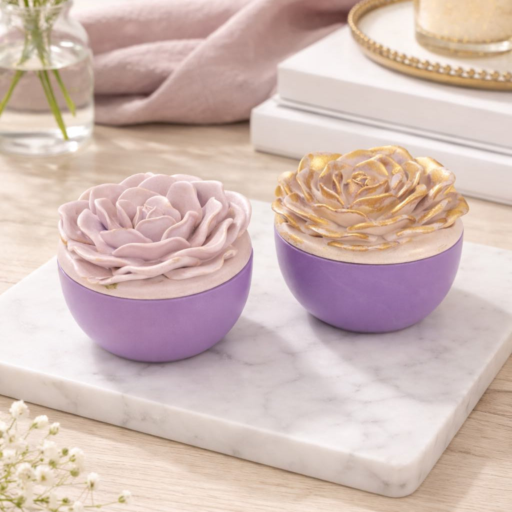 Two decorative items resembling flowers in purple bowls on a marble surface with a candle and flowers in the background.