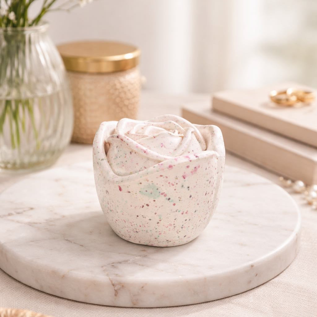 Small white ceramic pot with speckled design on a marble surface, with flowers and candles in the background.